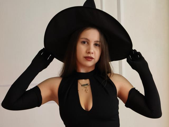 LilyMeeow with black witch hat, black sleeveless top with cutout, black gloves, holding hat, small tattoo under cleavage, brown hair, light skin, serious expression.
