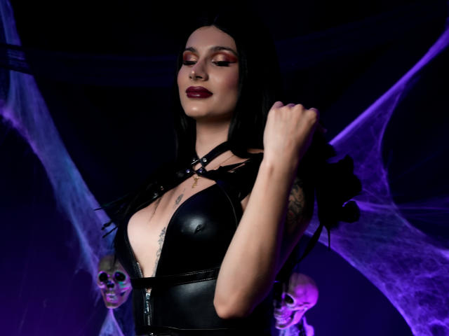 Maddymayer with black leather top, deep cleavage, large tits, dark lipstick, holding black feather, tattoo on chest, skulls in background, purple spider web, dark hair, confident expression.