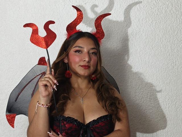 DuboisCarla with red glitter devil horns, black and red lace-up corset, red pom-pom earrings, and small grey bat wings, holding a red devil trident, her tits prominent.