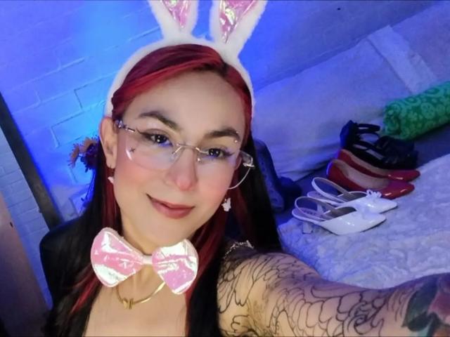 AmyStreetwise69 with red hair, glasses, bunny ears, pink bowtie, tattoos on arm, smiling, wearing makeup, in a blue-lit room, holding white heels, and red shoes.