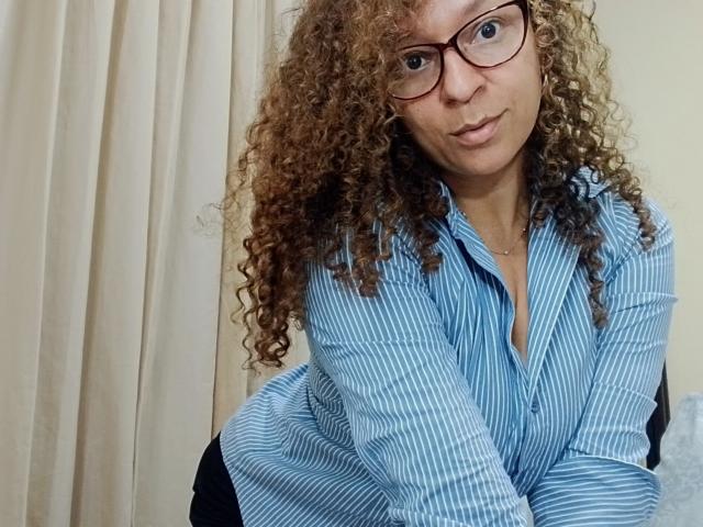 Angelonix with curly brown hair, wearing glasses and a blue striped shirt, leaning forward with a playful, slightly surprised expression, showing a bit of cleavage.