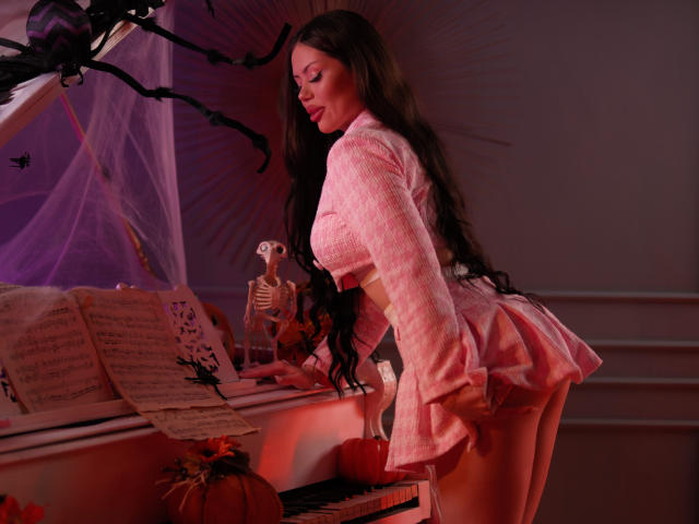 AlessiaNoire with long brown hair, pink plaid shirt, and pink mini-skirt bends over a piano, showing her ass and small white panties. Pumpkin and skeleton on piano.
