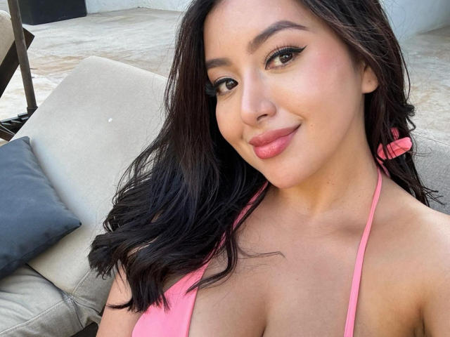 MsBunngy with dark wavy hair, pink bikini top, and cleavage, smiling at the camera, showing off her pink lips and eyeliner. Her tits are slightly visible.