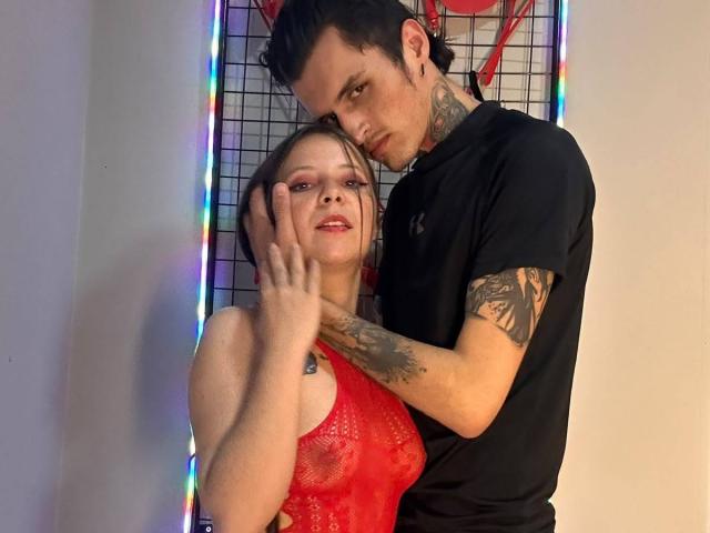 GraceAndTony with tattoos, black shirt, and short dark hair, leaning over GraceAndTony in red lace lingerie, touching her cheek, colorful lights in background. GraceAndTony's tits visible.