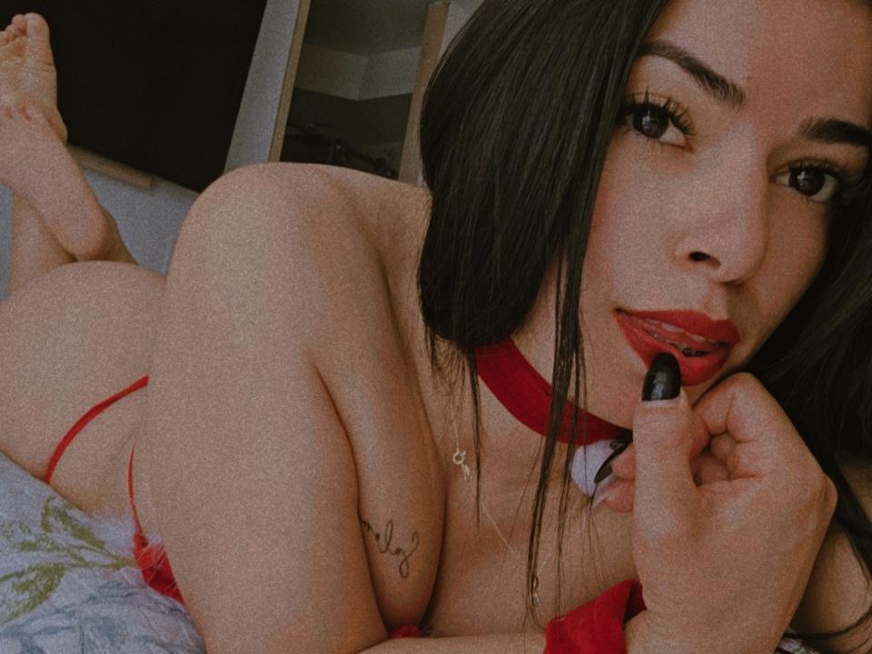 Steefany with long black hair, red lipstick, and black nail polish, lying on her stomach, wearing red lingerie, biting her finger, showing her ass, and a small tattoo on her arm.