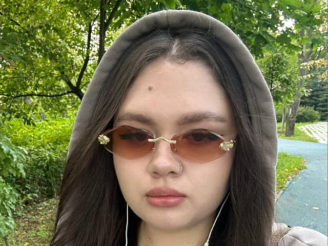 Corrine Preisser, avec ses yeux marron, son anneau de nez et son sweat à capuche marron, porte des lunettes de soleil rose avec des motifs floraux, ainsi que des écouteurs blancs, dans un parc verdoyant et ombragé..