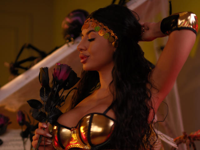 LatinaLoco with big boobs, gold armor, black hair, and a golden headpiece, holding a rose, eyes closed, arm raised, in a warm, yellow-lit room.
