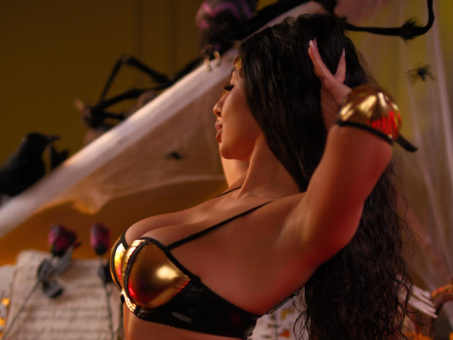 LatinaLoco with long dark hair, gold and black metallic bra showing big tits, arms raised, wearing gold bracelets, side profile, soft light, slight red glow, sensual pose.