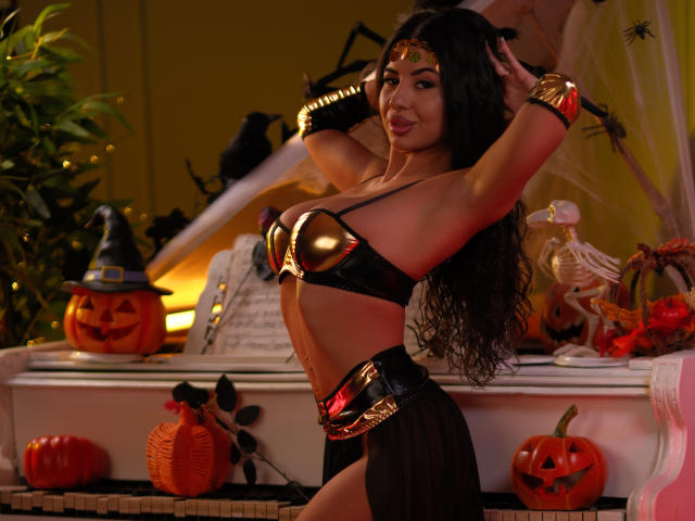 LatinaLoco with long black hair, wearing a gold and black Wonder Woman-style bikini, gold headpiece, and bracelets, posing with hands in hair, showing her tits and ass.
