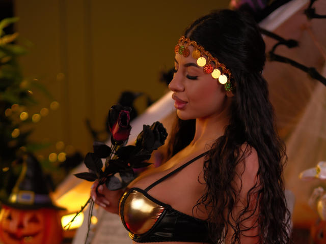 LatinaLoco with curly black hair, wearing a gold headband and black leather top, holding black roses, showing her big boobs and gold nipple cover, in a Halloween-themed setting.