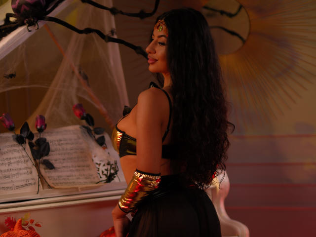 LatinLoco with long black curly hair, gold metallic top, black sheer skirt, gold arm bracelet, looking back, showing her ass, in a dimly lit, warm-toned room.