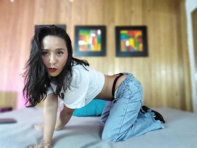 AgathaTeranh on all fours on a bed, wearing a white crop top, blue jeans, black underwear, and red lipstick, with long black hair and a wooden wall behind her.