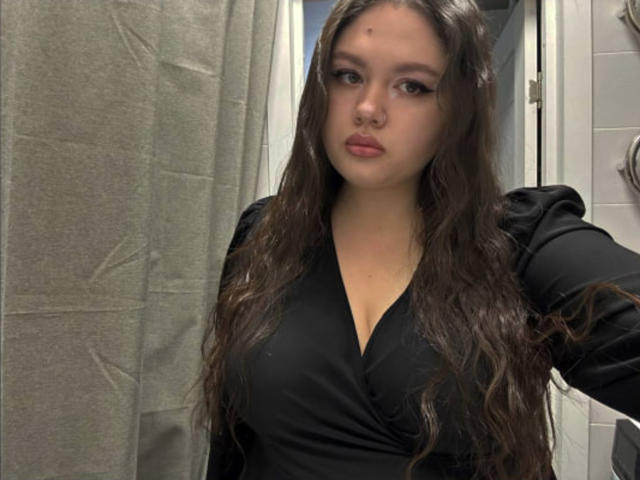 Corrine Preisser, con cabello castaño ondulado largo, vestida con una blusa negra de cuello en V, con mangas negras abullonadas y un piercing en la nariz, con senos pequeños, mirando a la cámara con una expresión neutra..