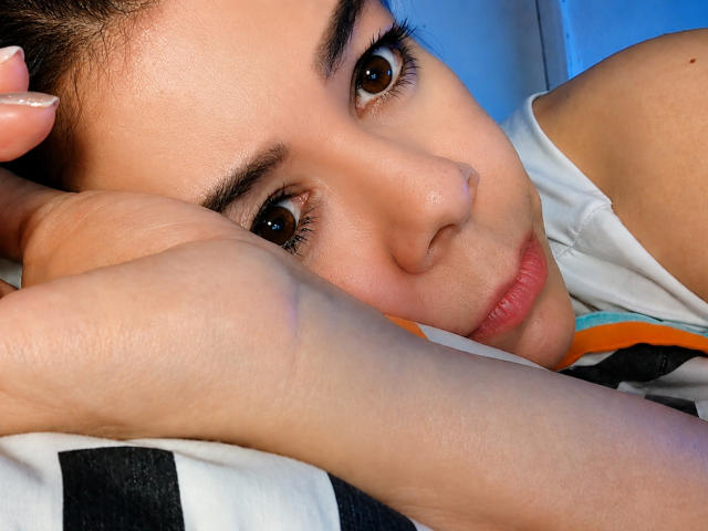 EmiliaMilf's face, brown eyes, and slightly parted lips with a hand resting on her cheek, wearing a white shirt, has a soft, relaxed expression, and light skin.