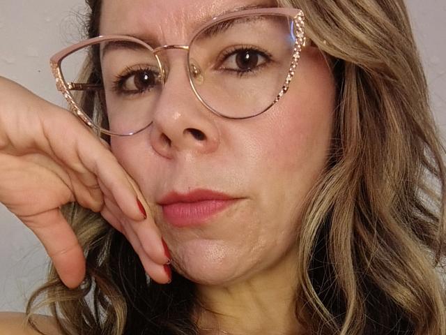 EmiliaMilf has a pensive look with pink-tinted glasses, wavy brown hair, red nails, and slightly shiny skin. She's touching her cheek with her left hand.