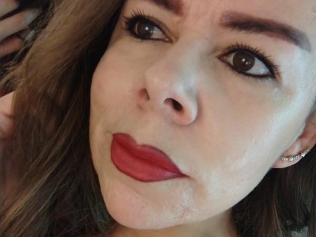 EmiliaMilf's face with puffy red lipstick, dark eyeliner, and slightly upturned nose, showing a subtle smile. She has brown eyes and wavy blonde hair, wearing small earrings.