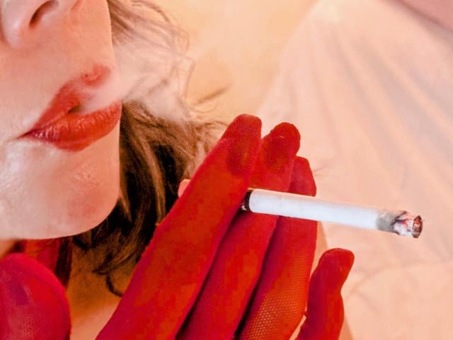 EmiliaMilf with red gloves, puffy red lips, and a cigarette, exhaling smoke. Her face is partially visible, with wavy brown hair. The cigarette has a burned tip.
