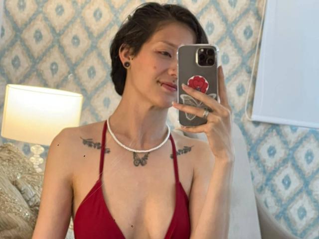 LissyLuxe, com cabelo castanho curto, seios pequenos, top vermelho de alças, alargadores de orelha pretos, tatuagens nos ombros, sorrindo, segurando um celular com um suporte vermelho, em um quarto aconchegante..