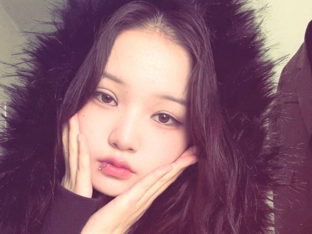 RayeAbend has pale skin, dark brown eyes, and a black fur hoodie. She's touching her face with both hands, showing a pierced lower lip. Her hair is straight and black.