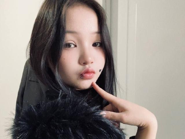 RayeAbend has black hair, a pierced lower lip, and a black mole on her cheek. She's pouting with her finger on her chin, wearing a black fur collar.