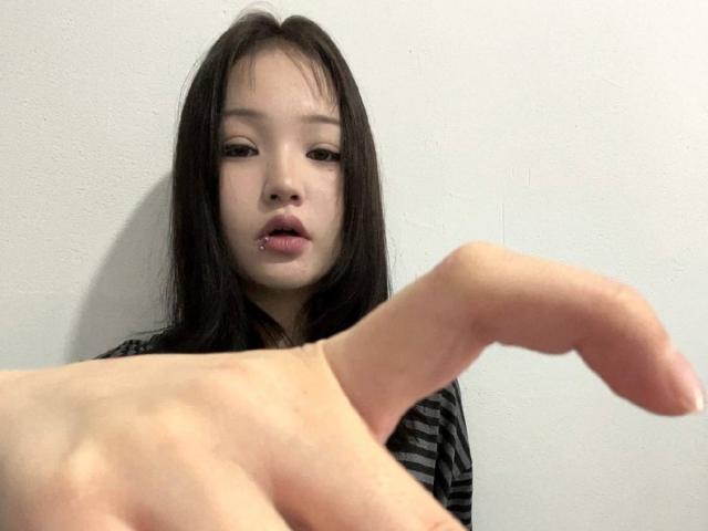 RayeAbend has black hair, a pierced lower lip, and a serious expression. Her hand is in the foreground, slightly out of focus. She wears a striped shirt.