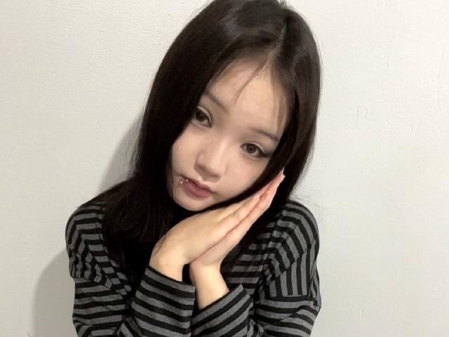 RayeAbend with black hair, striped shirt, hands on face, looking down, slight pout, small nose piercing, white wall, soft light, subtle makeup, slight sadness in eyes.