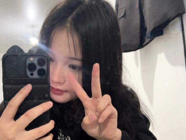 RayeAbend with black hair, taking a selfie in a mirror, making a peace sign with her right hand, wearing a black top, holding a black phone with a flash.