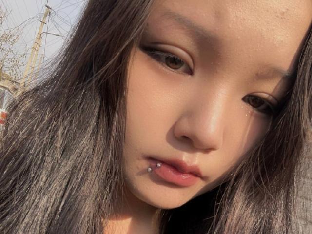 RayeAbend has a pouty face with a lip piercing on her bottom lip, straight black hair, and brown eyes. Her skin is light with a slight sheen.