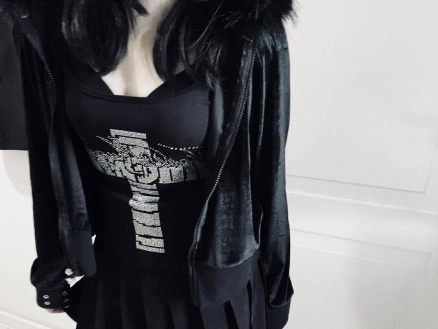 RayeAbend with black hair, wearing a black cross shirt, black dress, and a shiny black jacket with fur trim, has visible tits and a black belt with studs.