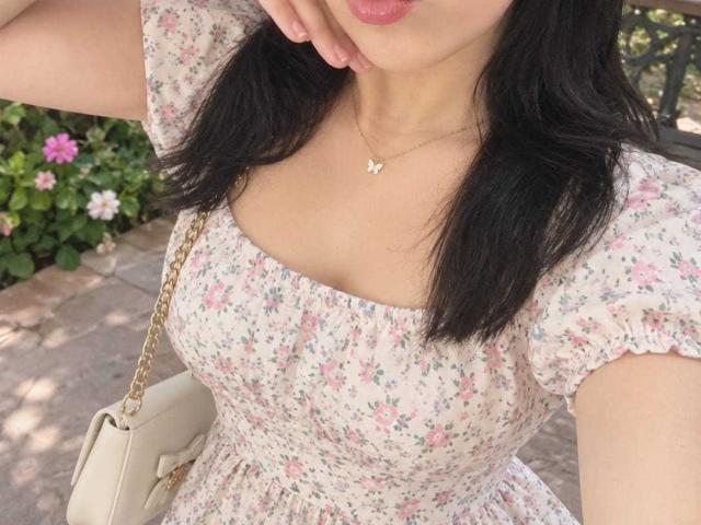 NenaBlack with dark hair, wearing a floral dress, her tits showing under the fabric, white purse on shoulder, gold chain necklace with butterfly pendant, pink lipstick, hand by mouth.