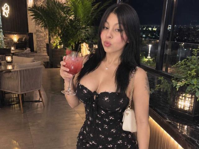 NenaBlack with black floral dress, holding red cocktail, large boobs, long black hair, pouty lips, white purse, black nails, standing in dimly lit room, night city view.