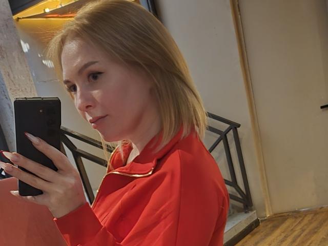 KatrinaSi con i capelli biondi, indossa una camicia rossa con zip, tiene in mano un telefono nero, sta scattando un selfie, guarda verso la fotocamera, in una stanza poco illuminata, pavimento in legno, ringhiera metallica sullo sfondo..