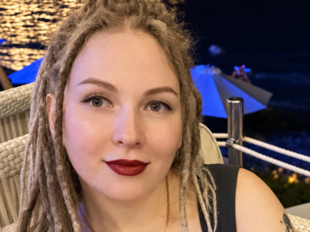 KatrinaSi, avec ses dreadlocks blondes, son rouge à lèvres rouge et ses yeux verts, regarde l'appareil photo, assise à l'extérieur la nuit, vêtue d'un haut noir et d'un collier en or..