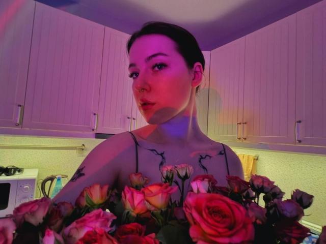 DeneenAngelone with tattoos, holding a bouquet of pink and red roses, has short dark hair, and a nose ring, illuminated by purple and pink lighting.