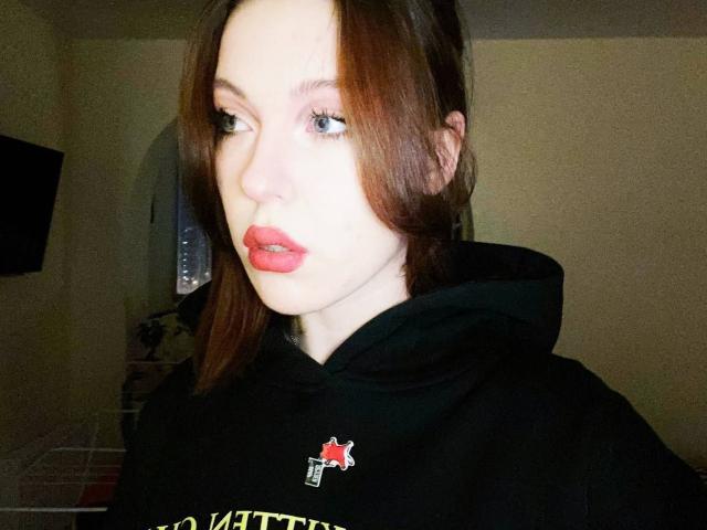 DeneenAngelone has pale skin, red lipstick, and brown hair. She's wearing a black hoodie with a red and yellow emblem, looking to the side with blue eyes.