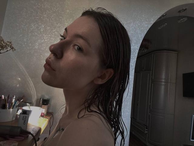DeneenAngelone with wet brown hair, pouty lips, and a slight nose ring, wearing a sleeveless top, showing a tattoo on her shoulder, in a dimly lit room.