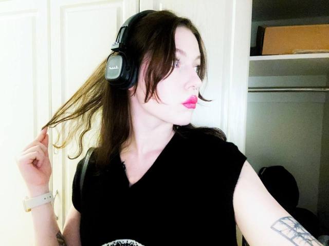 DeneenAngelone with black headphones, holding brown hair, wearing a black shirt, pink lipstick, white watch, and tattoo on right arm, looking to the side.