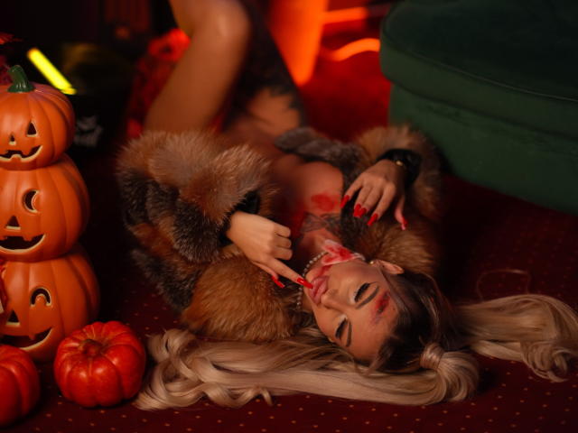 CelineDian, lying on red carpet, wearing fur jacket, biting red apple with blood, long blonde hair, green nails, surrounded by jack-o'-lanterns, green cushion, and small pumpkins.