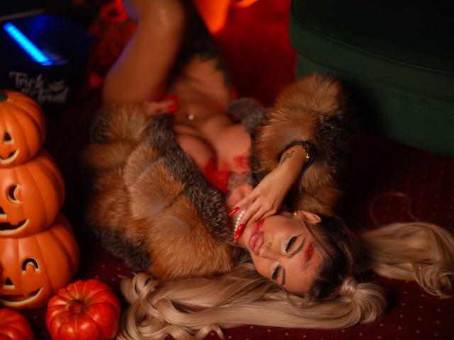 CelineDian with long blonde hair, lying on red carpet, wearing furry coat, red lipstick, touching her red-manicured nails, surrounded by jack-o'-lanterns, showing her tits and ass.