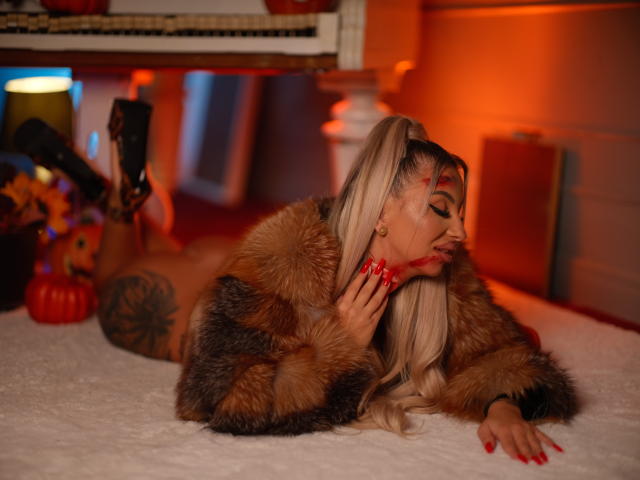 CelineDian lies on a bed, wearing a brown fur coat, red nails, and black high heels, with a tattoo on her thigh, and blonde hair in a ponytail.
