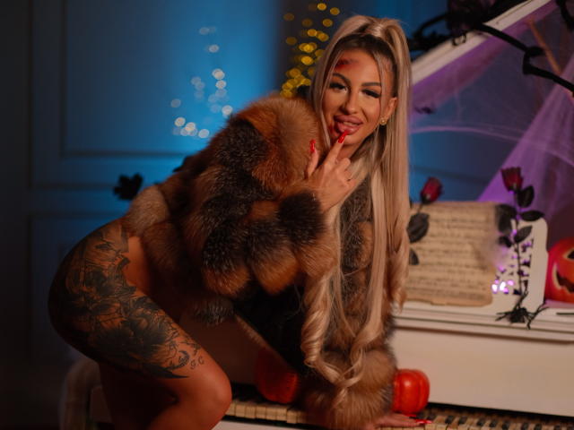 CelineDian with long blonde hair, wearing a fur coat, touching her red-lipped, smiling face, showing a tattoo on her left thigh, and a red apple on a piano.