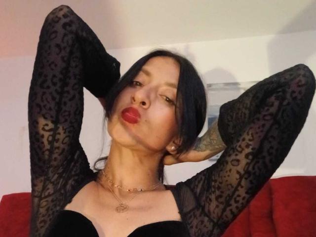 ZarayFeet with black lace sleeves, arms behind head, pouty red lips, tattooed arm, gold necklace, black bra, and red couch in background. Her tits are small.