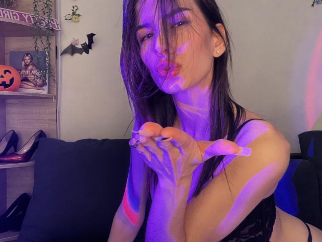 Valnes, con el pelo largo y castaño, vistiendo una blusa de encaje negro, labios fruncidos, un brillo púrpura y rosa en la piel, manos cerca de la cara, luz púrpura, mostrando sus pequeños senos, sentada en un sofá..