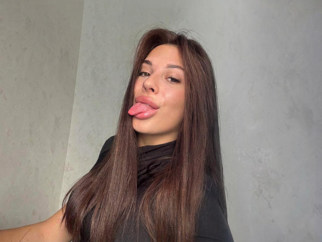 BlackFatal with long brown hair, sticking out her pink tongue, wearing a black shirt, has a slightly smug expression, and is looking at the camera.