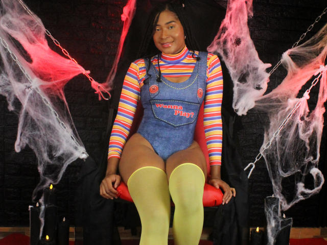 NaomiDaviss, with brown skin and long black hair, sits on a red chair, wearing a blue denim onesie, rainbow striped shirt, and yellow thigh-highs, surrounded by smoke.