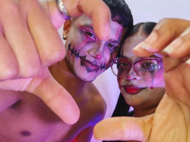 VanyAngel and VanyAngel, shirtless with makeup, showing off their faces with purple and black designs, and holding up their fingers, wearing glasses and red lipstick.