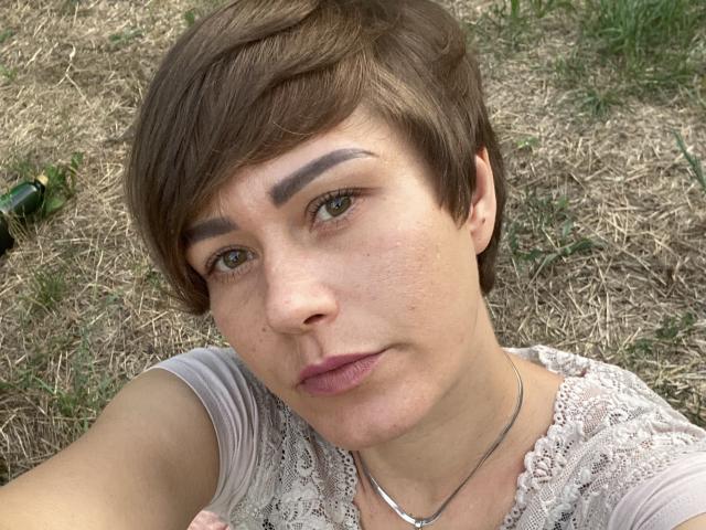 LenaShella has short brown hair, green eyes, and a serious expression. She's wearing a lace top and a silver necklace, with a slightly flushed face and pink lips.