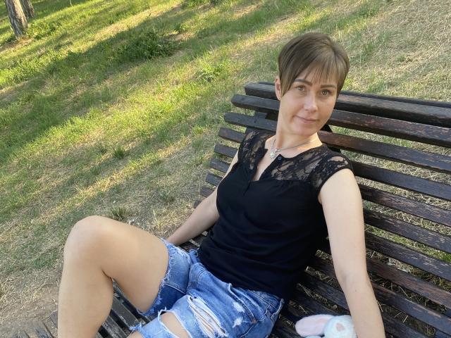 LenaShella, con i capelli castano corti, una maglia di pizzo nera e pantaloncini di jeans strappati, seduta su una panchina di legno, con le gambe incrociate, seno piccolo, carina, rilassata, con un leggero sorriso, in un parco con erba..