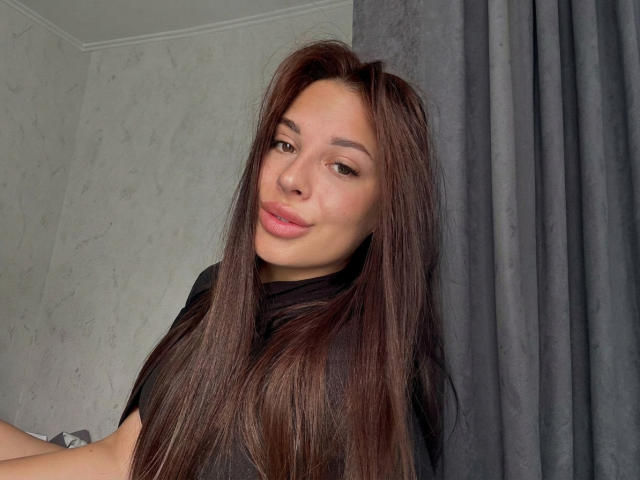 BlackFatal with long brown hair, light skin, and pink lips, wearing a black shirt, looking at the camera with a slight smile, standing in front of gray curtains.
