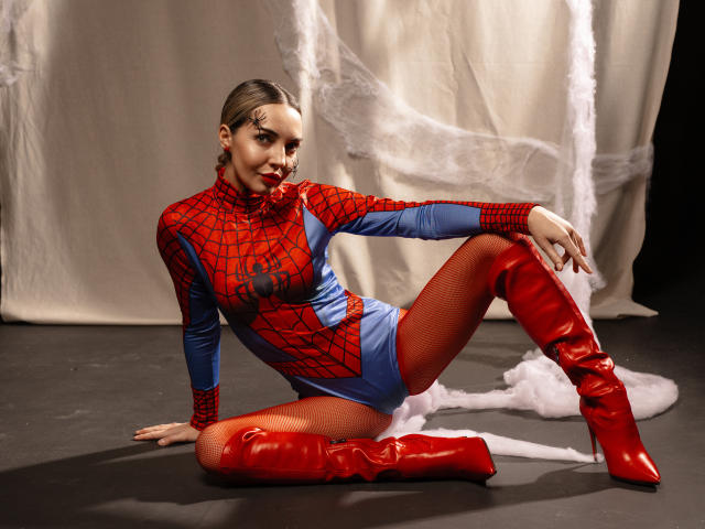 LettieSweet in a Spider-Man costume, red boots, and fishnet tights, sitting on the floor, legs crossed, arms resting, smiling, with white smoke on the ground.